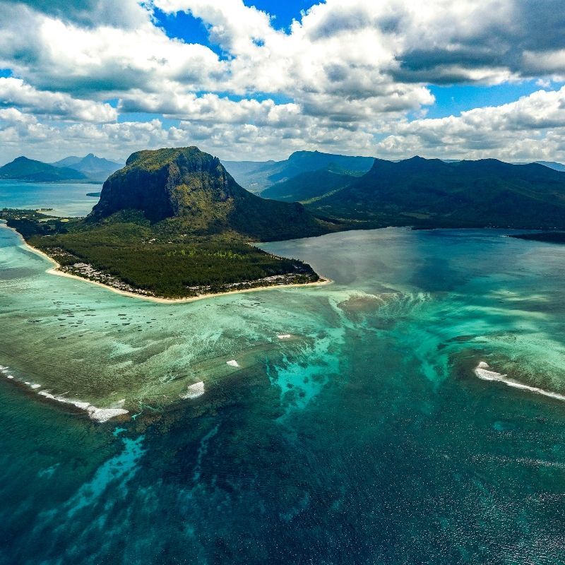 An aerial view captures the dramatic Le Morne Brabant Peninsula in Mauritius, rising majestically from the turquoise waters of the Indian Ocean, in this breathtaking travel photograph. The rugged mountain, a UNESCO World Heritage Site, is surrounded by pristine beaches, coral reefs, and the vibrant, shallow lagoons that shimmer in shades of blue and green. Le Morne is renowned for its natural beauty, cultural history, and recreational opportunities, including hiking, snorkeling, and kite surfing. Ideal for photographers, adventure travelers, and nature enthusiasts, this scene showcases the striking contrast between mountain, sea, and sand in one of Mauritius’ most iconic landscapes. Inspiration Africa specializes in bespoke, tailor-made journeys to Mauritius and across Africa, crafting unforgettable coastal and mountain experiences in extraordinary destinations.