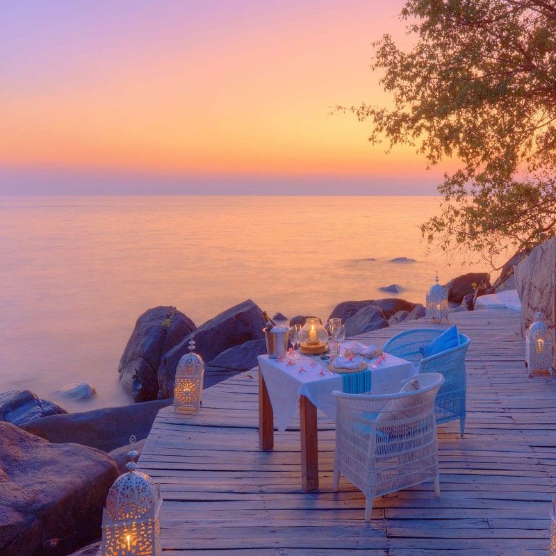 Outdoor dining on the veranda at Kaya Mawa in Malawi, set beside the tranquil waters of Lake Malawi at sunset, is captured in this evocative travel photograph. Tables are elegantly arranged to face the lake, where golden light reflects across the calm surface and the sky shifts through warm hues of orange and pink. The open-air setting blends seamlessly with the surrounding gardens and shoreline, creating an atmosphere of intimacy, relaxation, and natural beauty. Soft evening light enhances the textures and colors of the scene, emphasizing the lodge’s harmonious lakeside design. Inspiration Africa curates bespoke, tailor-made journeys to Malawi and across Africa, combining luxury accommodations with immersive wildlife, cultural, and scenic experiences.