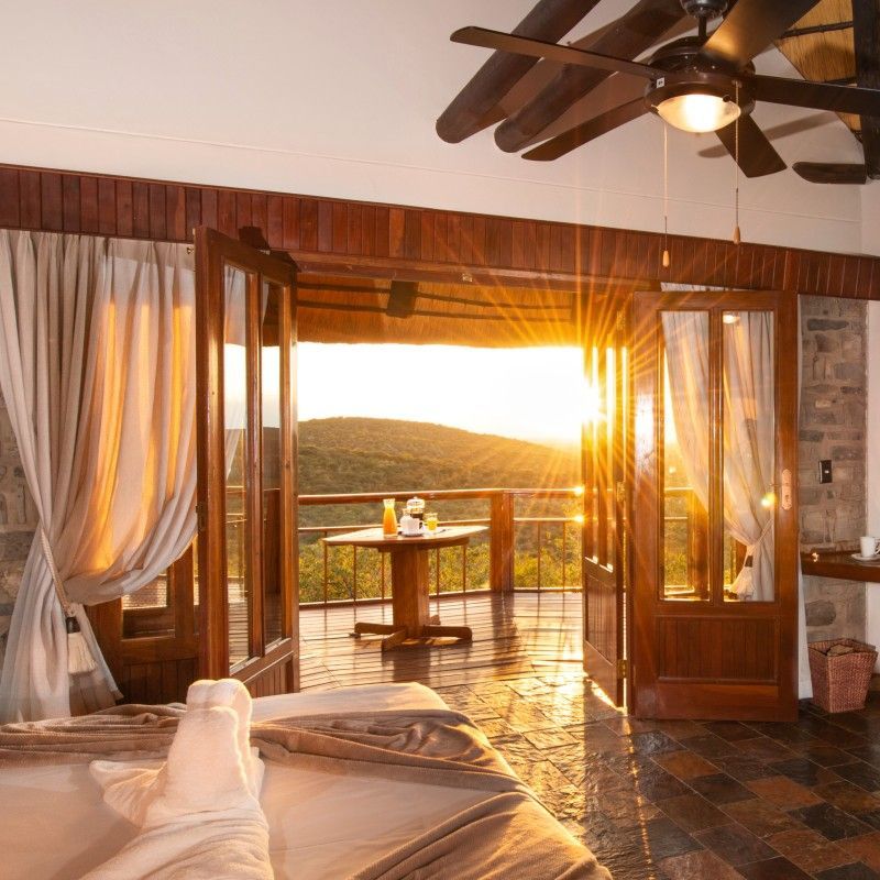 One of the beautifully designed rooms at Etosha Mountain Lodge in Namibia is captured at sunset in this atmospheric travel photograph, bathed in warm golden light. As the sun dips below the horizon, soft hues of orange and amber filter through the windows, illuminating the elegant interiors and enhancing the sense of calm and comfort. The room opens onto sweeping views of the surrounding plains, where the changing sky creates a dramatic backdrop to the lodge’s elevated setting. This sunset moment perfectly reflects the lodge’s blend of refined comfort and wilderness immersion, offering guests a peaceful retreat after a day of safari exploration. Ideal for luxury travelers and safari enthusiasts, this scene captures the romance, tranquility, and timeless beauty of Etosha Mountain Lodge. Inspiration Africa specializes in bespoke, tailor-made journeys to Namibia and across Africa, crafting unforgettable safari stays and immersive lodge experiences in extraordinary destinations.