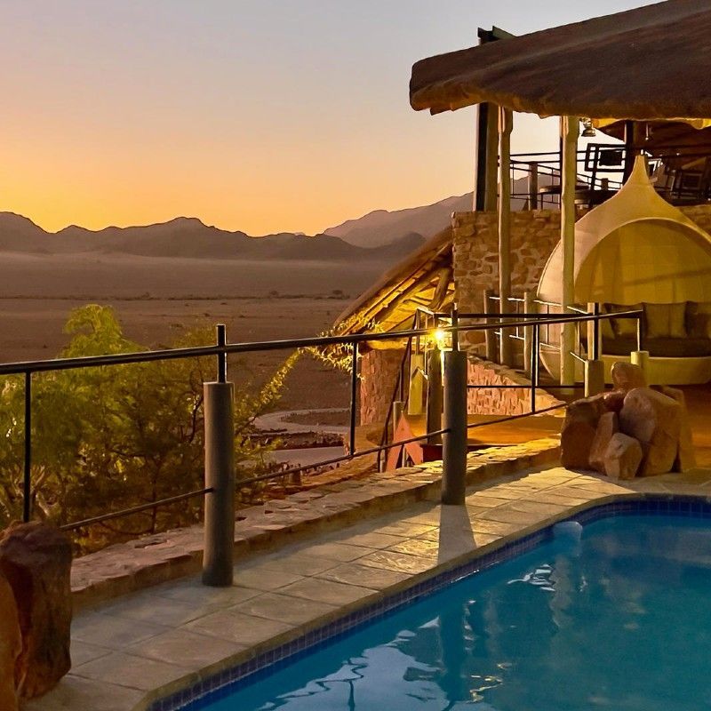 The swimming pool at Namib Outpost in Namibia is captured in this striking travel photograph, set against the dramatic backdrop of the vast Namib Desert and rugged mountains. The pool’s calm waters reflect the clear blue sky, offering a refreshing oasis amid the arid landscape. Sun loungers and shaded seating provide spaces for relaxation while taking in the sweeping desert vistas, and the natural, minimalist design blends seamlessly with the surrounding environment. The scene conveys tranquility, adventure, and understated luxury, highlighting Namib Outpost as a serene retreat where guests can unwind while fully immersed in Namibia’s iconic desert scenery. Inspiration Africa specializes in bespoke, tailor-made journeys to Namibia and across Africa, crafting unforgettable travel experiences in extraordinary destinations.