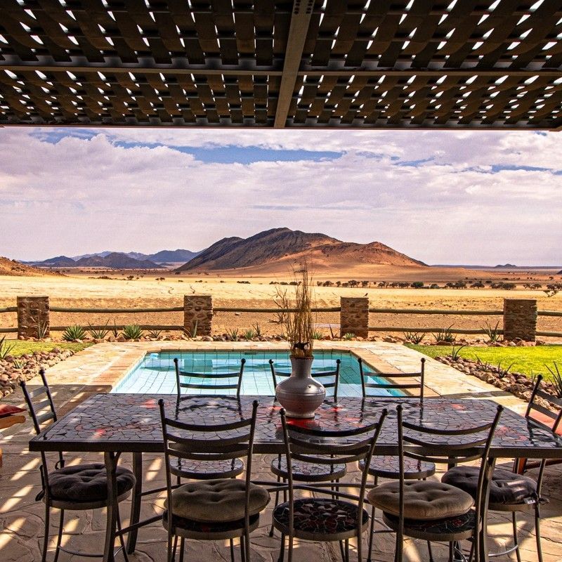 The outdoor dining table and chairs on the patio of Greenfire Desert Lodge in Namibia are captured in this inviting travel photograph, set against the vast, open beauty of the desert landscape. Thoughtfully arranged furnishings blend natural materials with understated elegance, creating a relaxed yet refined space for open-air dining. From the patio, sweeping views stretch across the surrounding plains, where changing light and endless horizons enhance every meal with a sense of place and tranquility. This outdoor setting reflects the lodge’s intimate, eco-conscious philosophy, offering guests a chance to connect deeply with nature while enjoying comfort and simplicity. Ideal for travelers seeking authentic desert experiences and relaxed luxury, this scene captures the quiet charm and immersive atmosphere that define Greenfire Desert Lodge. Inspiration Africa specializes in bespoke, tailor-made journeys to Namibia and across Africa, crafting unforgettable lodge stays and immersive desert moments in extraordinary settings.