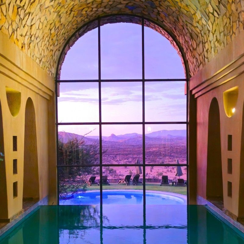 An elegant indoor swimming pool in the spa and wellness area of GocheGanas in Namibia is captured in this serene luxury travel photograph. A large glass window opens the space to sweeping views of the surrounding savannah, allowing natural light to flood the pool area and creating a calming connection between indoors and outdoors. The tranquil water, clean architectural lines, and minimalist design evoke a sense of relaxation and rejuvenation, perfectly complementing the lodge’s natural setting. This wellness space offers a peaceful retreat after days of exploration, where comfort, nature, and wellbeing come together. Ideal for luxury travelers and wellness enthusiasts, this scene highlights the refined spa experience and quiet indulgence that define GocheGanas. Inspiration Africa specializes in bespoke, tailor-made journeys to Namibia and across Africa, crafting unforgettable lodge stays and restorative wellness experiences in extraordinary landscapes.