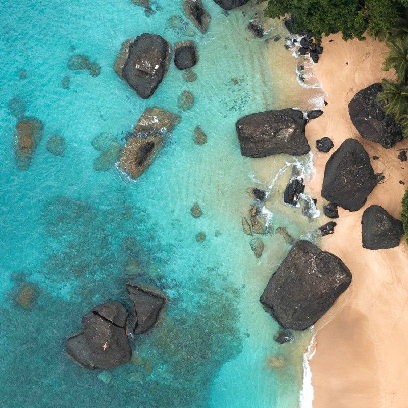 An aerial view of a secluded beach in São Tomé and Príncipe is captured in this breathtaking travel photograph, showcasing the island nation’s pristine coastline and natural beauty. Turquoise ocean waters gently meet soft sandy shores, while dark volcanic rocks and lush tropical vegetation frame the beach, creating a striking contrast of colors and textures. Sunlight reflects across the clear water, emphasizing the tranquility and untouched character of this remote island setting. The scene conveys serenity, escape, and the rich natural landscapes that define São Tomé and Príncipe. Inspiration Africa curates bespoke, tailor-made journeys to São Tomé and Príncipe and across Africa, combining immersive island experiences with wildlife, culture, and scenic exploration in extraordinary destinations.
