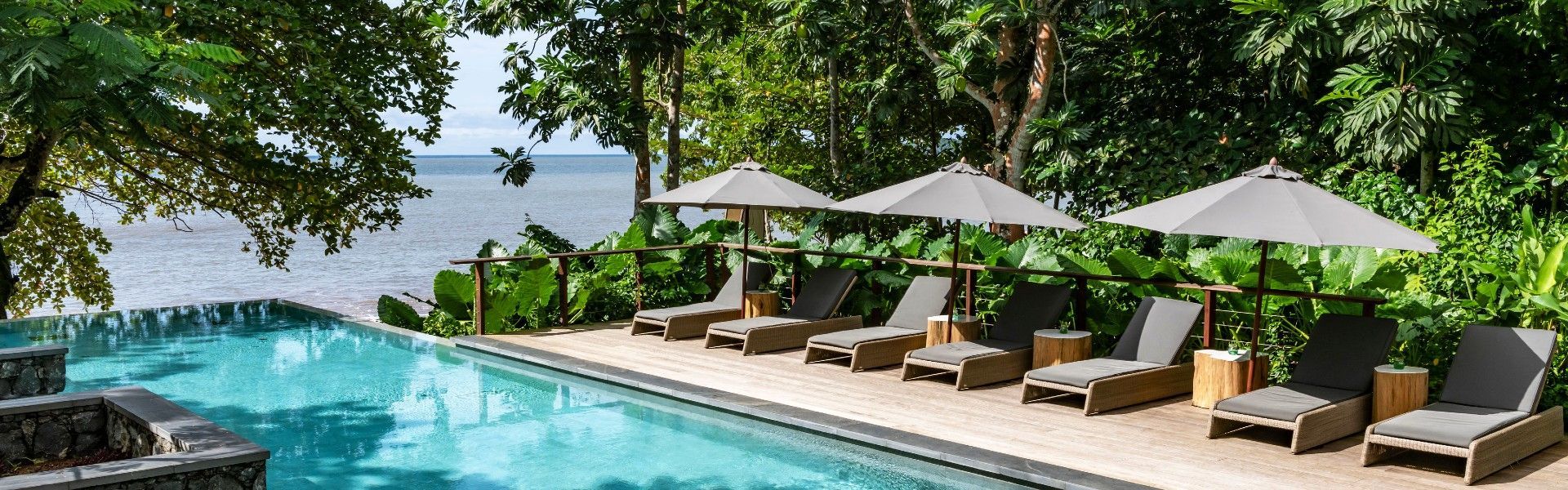 The oceanfront swimming pool at Sundy Praia in São Tomé and Príncipe is captured in this striking travel photograph, showcasing a secluded luxury retreat set between lush tropical forest and the Atlantic Ocean. The pool blends seamlessly into the natural landscape, framed by palm trees, soft sandy tones, and verdant greenery, with uninterrupted views toward the sea beyond. Sun loungers and shaded areas invite quiet relaxation, while the design reflects a balance of understated elegance and barefoot luxury. The scene conveys a sense of tranquility, exclusivity, and connection to nature, highlighting Sundy Praia as an idyllic island escape. Inspiration Africa specializes in bespoke, tailor-made journeys to São Tomé and Príncipe and across Africa, crafting unforgettable travel experiences in extraordinary destinations.