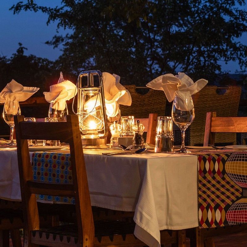 The exterior dining area at Masuwe Lodge in Zimbabwe is captured in this atmospheric nighttime travel photograph, showcasing a warm and inviting space illuminated by soft lighting. Tables are set outdoors, surrounded by lush vegetation and open skies, creating an intimate and tranquil ambiance for guests to enjoy meals under the stars. The subtle glow from lanterns and candles enhances the connection to the natural surroundings while providing a cozy and elegant atmosphere. The scene conveys the charm of dining in the African bush at night, combining comfort, scenic beauty, and the magic of Zimbabwe’s wilderness. Inspiration Africa specializes in bespoke, tailor-made journeys to Zimbabwe and across Africa, crafting unforgettable safari experiences in extraordinary destinations.