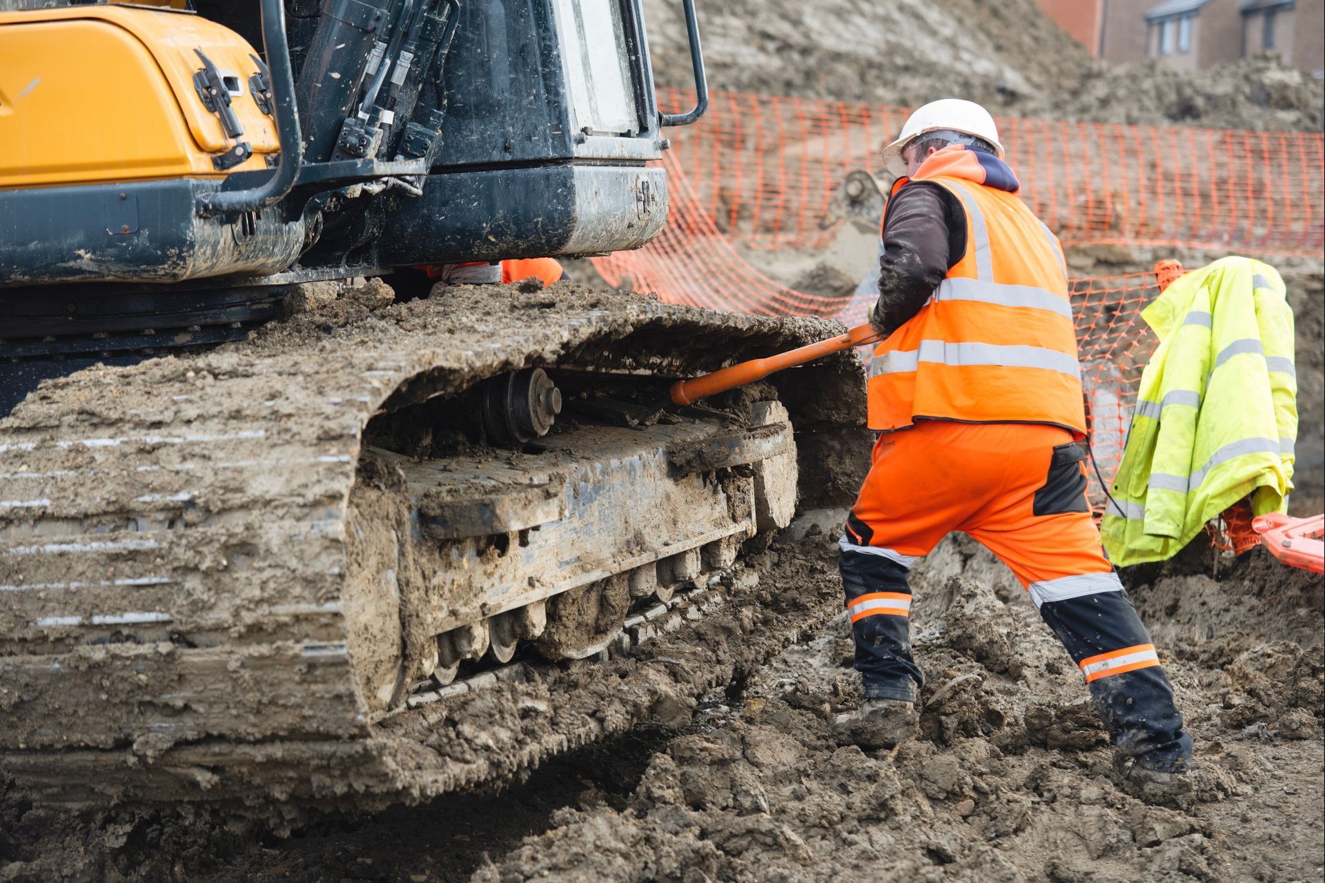 Construction worker clearing mud from the track of an excavator with a long-handled tool. Construction worker clearing mud from the track of an excavator with a long-handled tool.