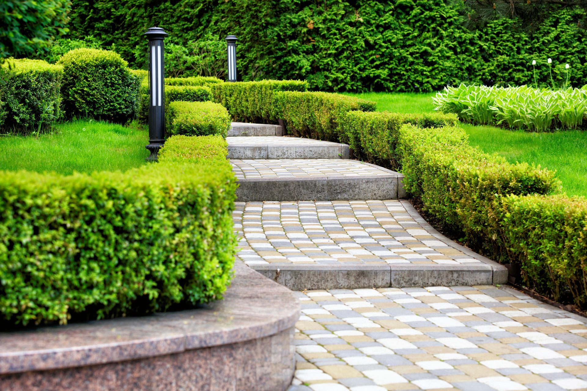 Stone pathway and steps ascending through manicured green hedges, with two black lamp posts.
