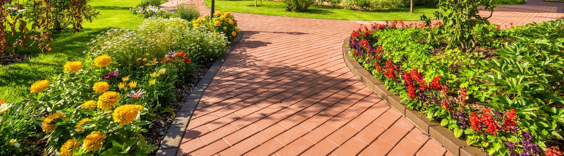 Brick pathway curves through a garden with flower beds and green grass.