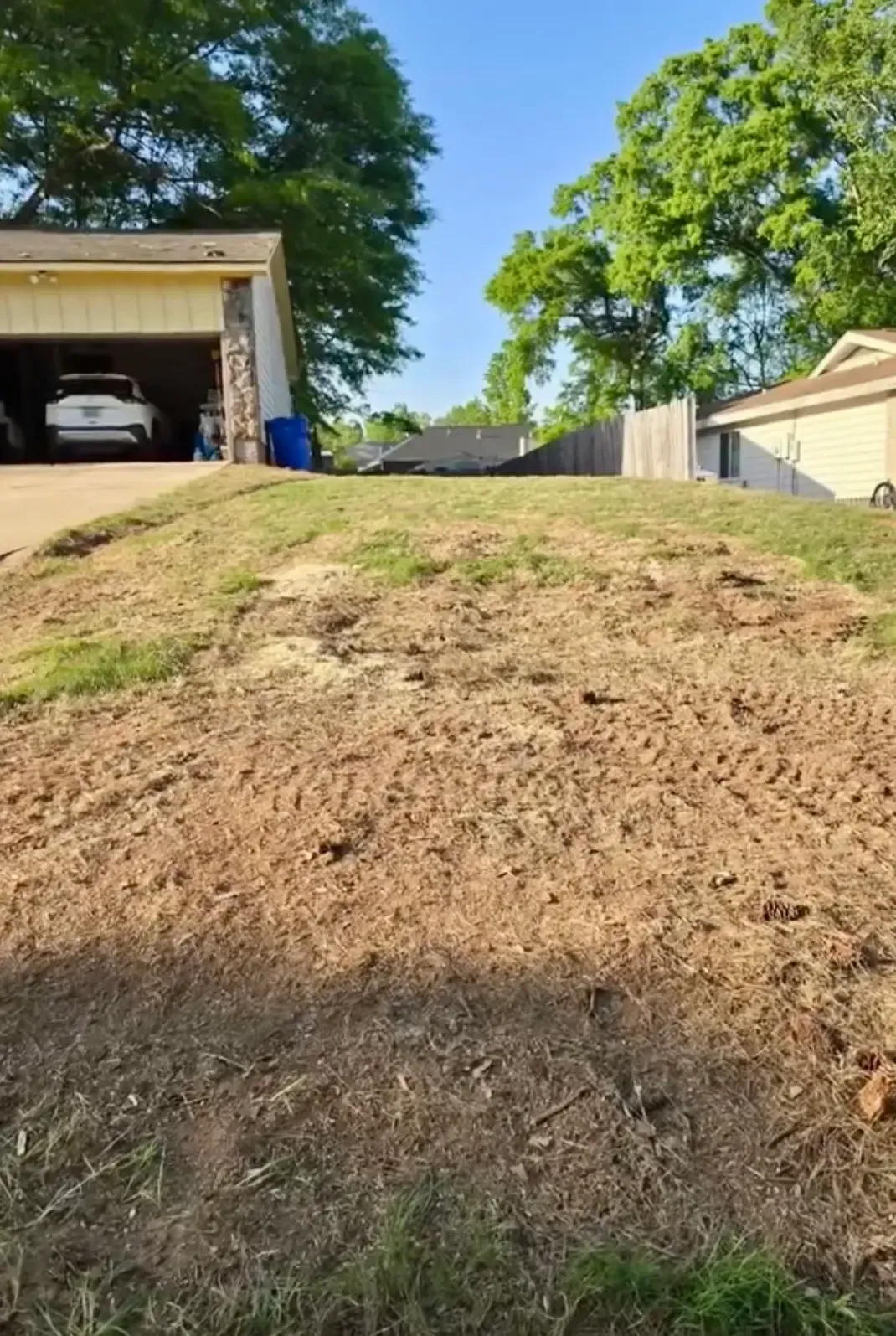 Bare dirt hill in a driveway beside garages and green trees on a sunny day