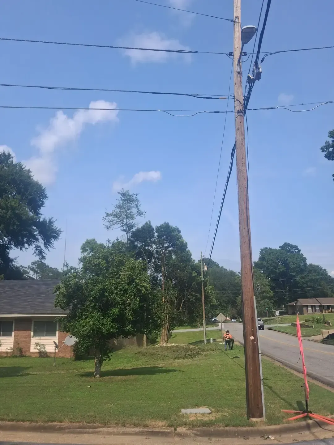 Residential street with utility pole, overhead power lines, trees, and a brick house under a blue sky.