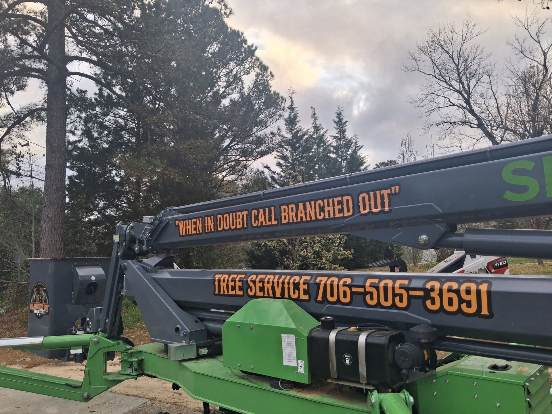 Green tree service lift truck with orange text parked outdoors near trees at dusk