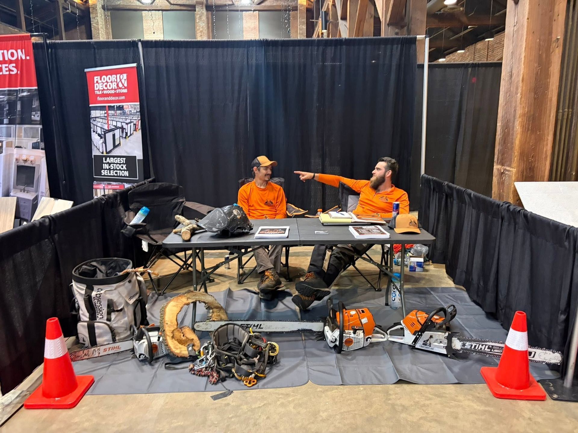 Two workers in orange shirts behind a booth with tools, cables, and traffic cones in a black-curtained display area