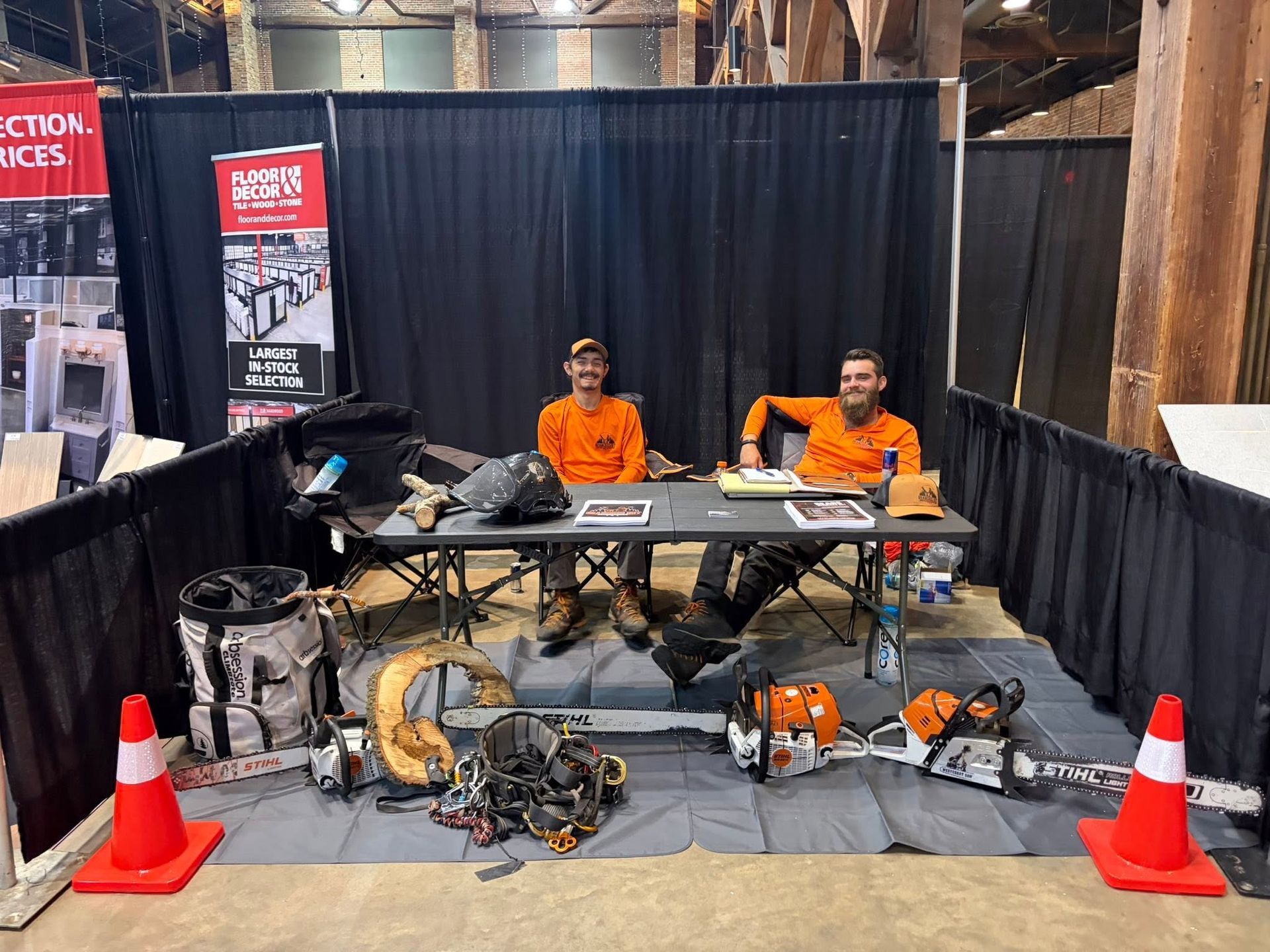 Two workers in orange shirts seated at a booth with tools and machinery displayed on a trade show floor