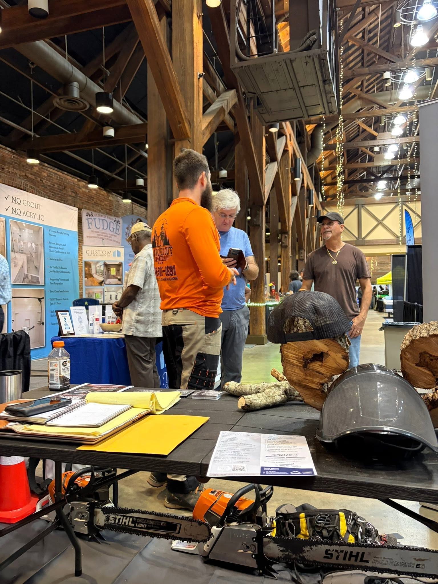 Indoor expo booth with people chatting beside a display table, posters, and a black helmet in a large hall