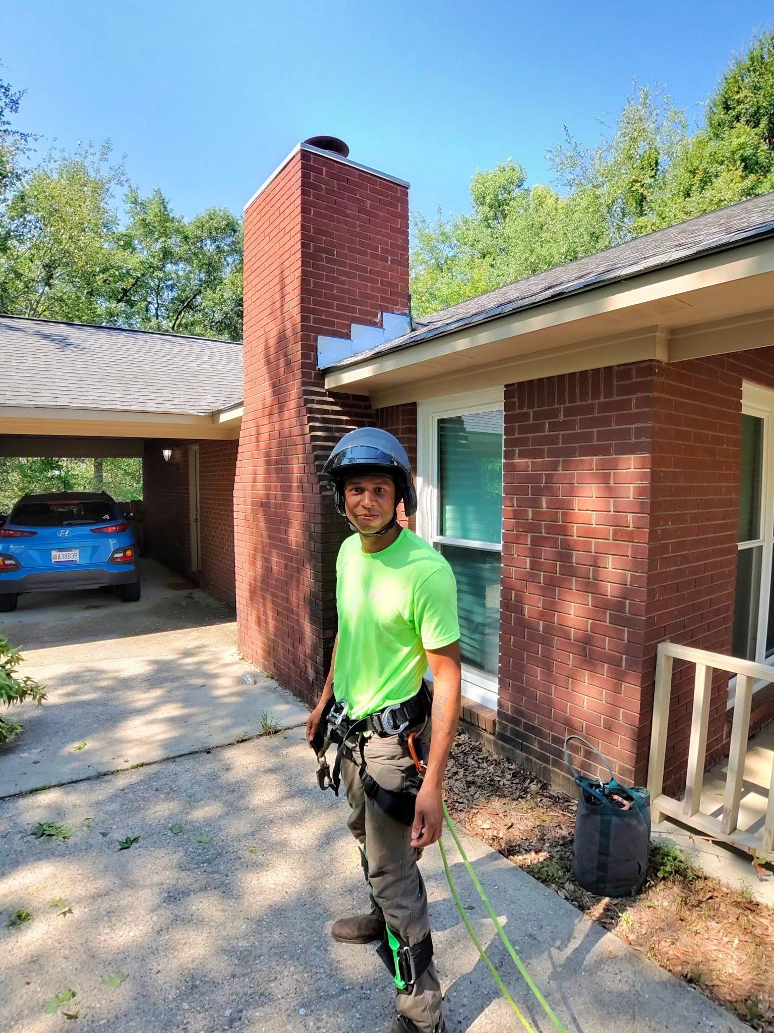 Person in neon green shirt and helmet standing outside a red brick house near a driveway and carport.