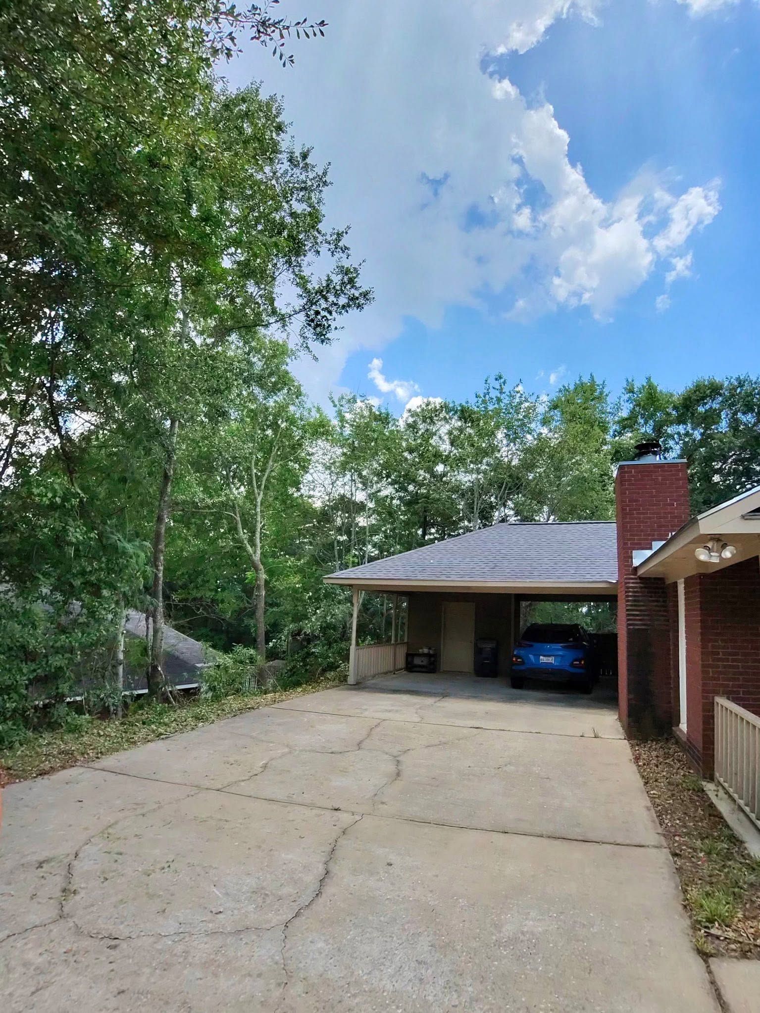Tree-lined driveway leading to a carport beside a brick house under a blue sky