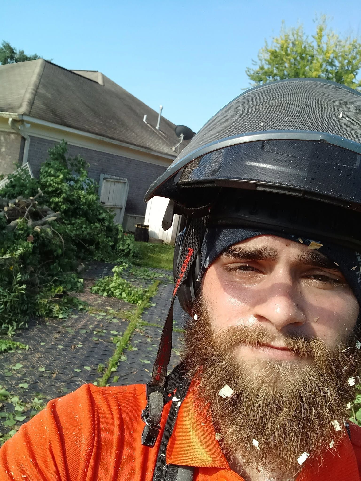 Bearded person in an orange shirt and black helmet taking a selfie outdoors by a house and greenery