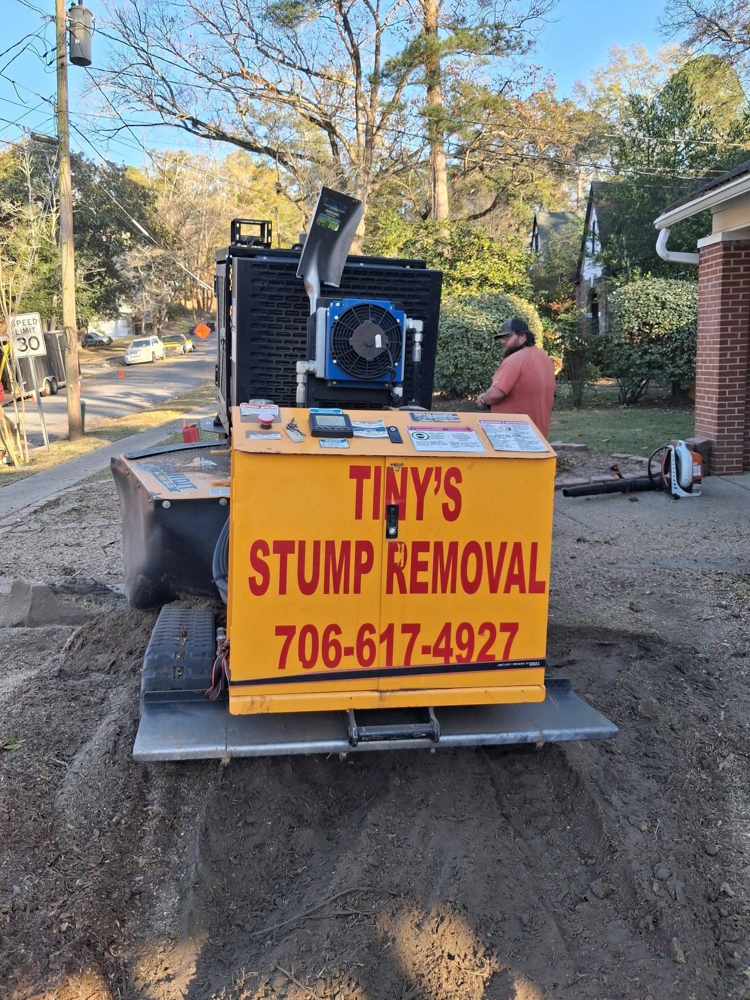 Yellow stump removal machine with “Tiny’s Tree Stump Removal” sign beside a large ground stump.