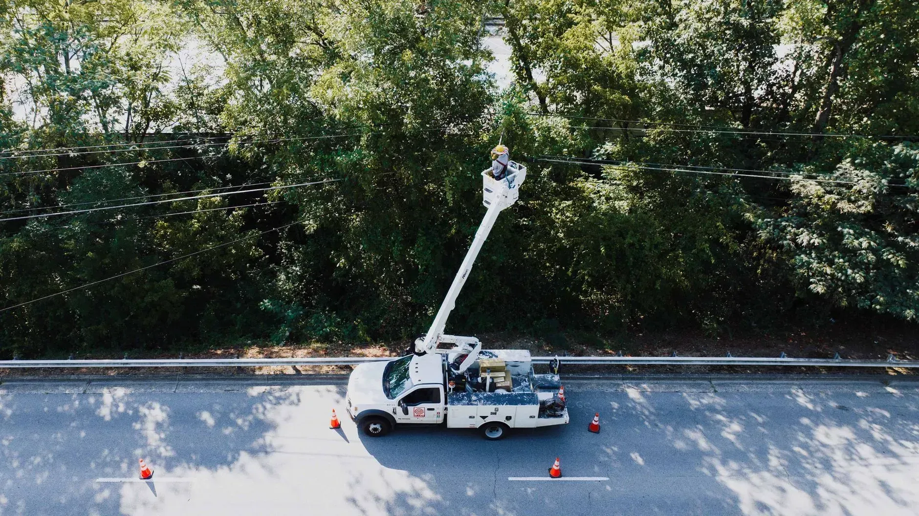 Bucket truck working by roadside beside trees and a guardrail, with cones marking the area.