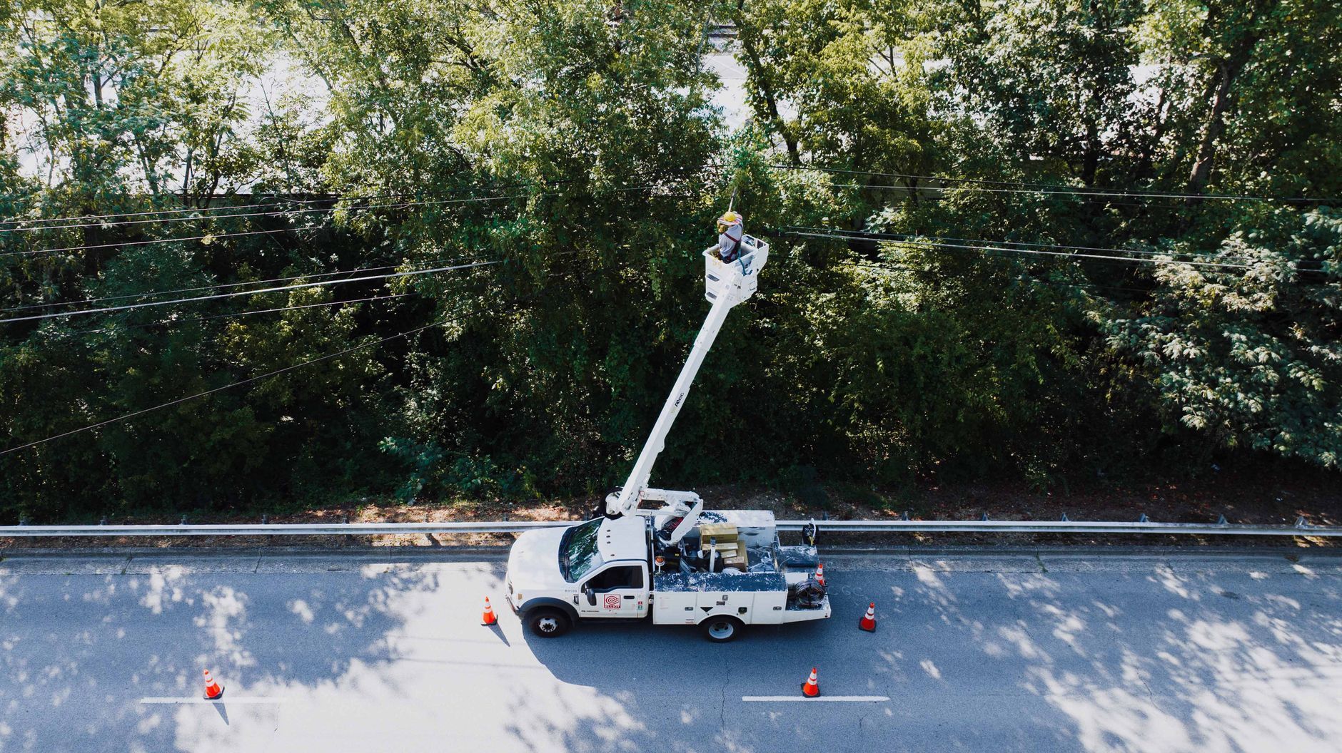 Bucket truck working by roadside beside trees and a guardrail, with cones marking the area.