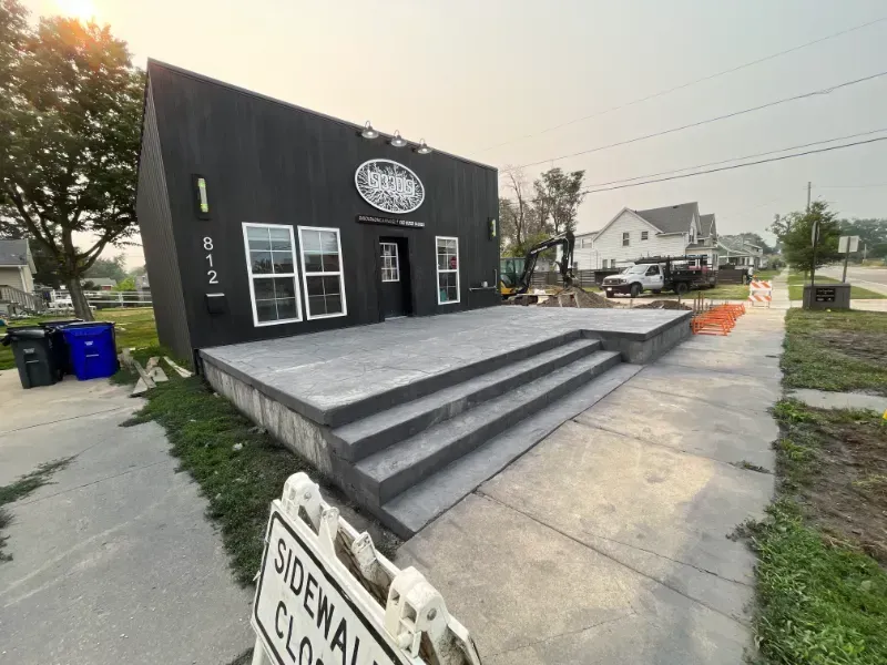 Small black storefront with gray steps and a circular sign, seen from a sidewalk on a cloudy day
