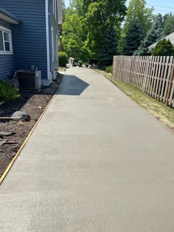 Fresh concrete driveway beside a blue house, with a wooden fence and trees along the side.