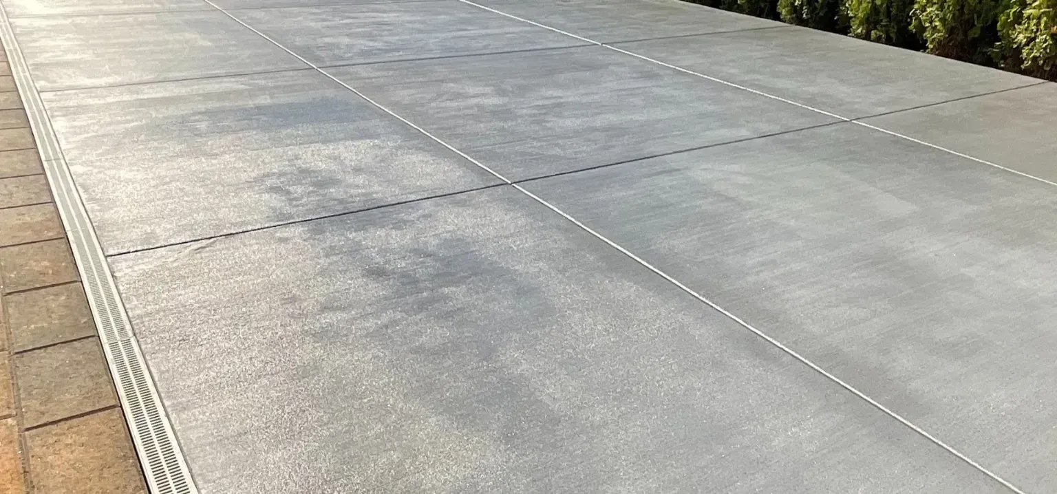 A clean, grey concrete driveway with evenly spaced control joints, bordered by pavers on the left and shrubs on the right.