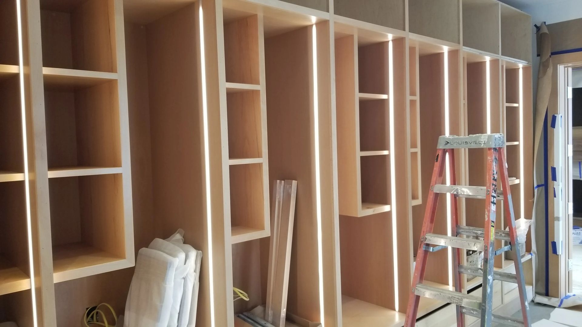 Row of unfinished wooden cabinets and shelving in a room, with a step ladder in front.