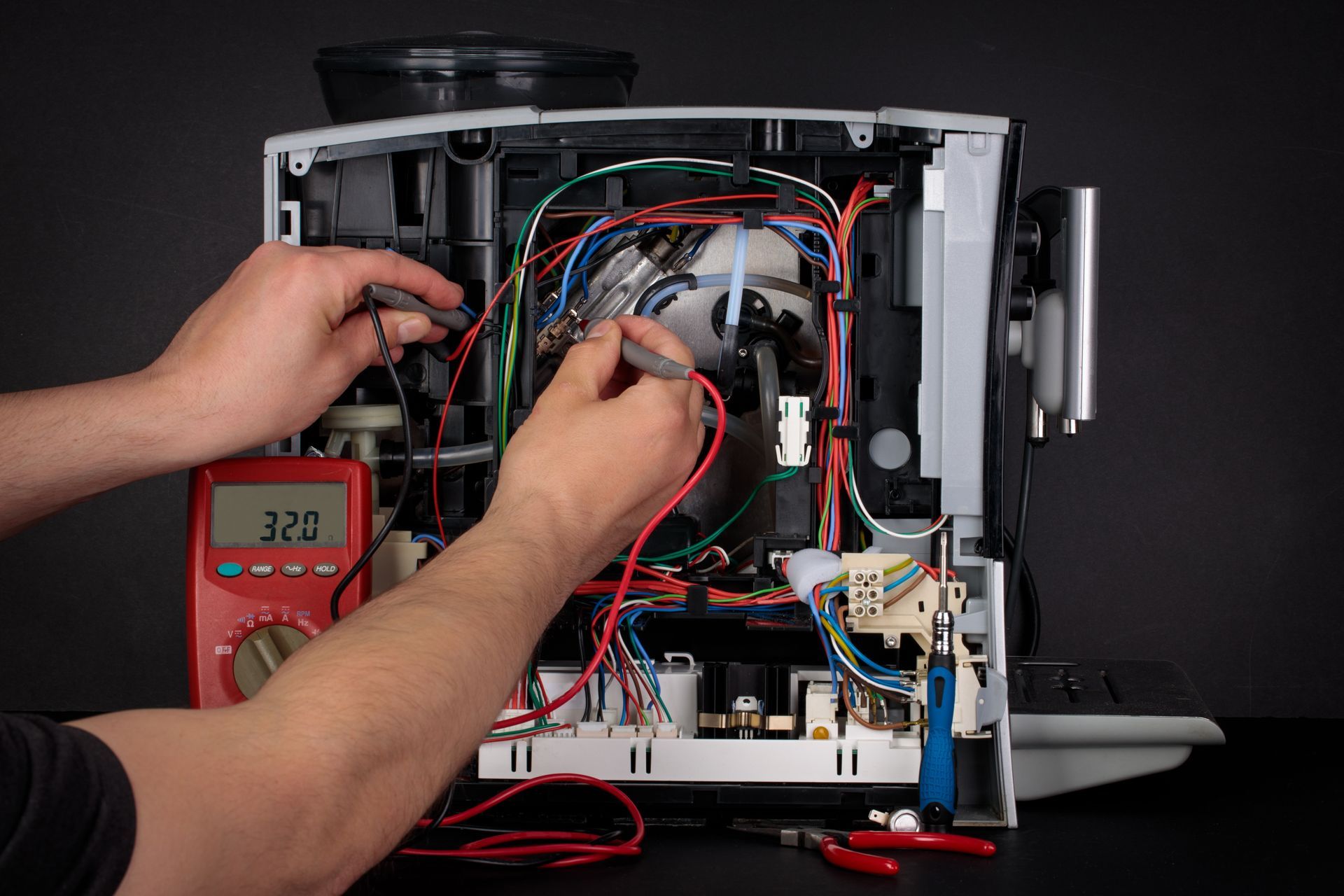Hands repairing wiring inside an open electronic device with a red multimeter beside it.