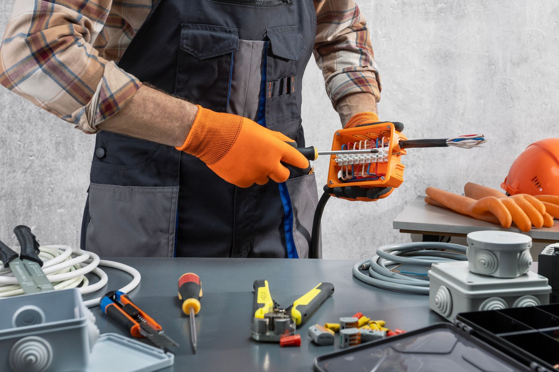 Worker using an orange tool to cut a cable at a workbench with tools and electrical parts