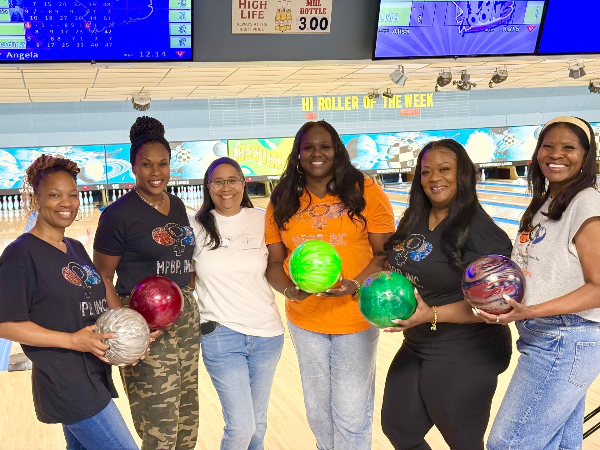 Six women at a bowling alley, smiling, holding bowling balls.