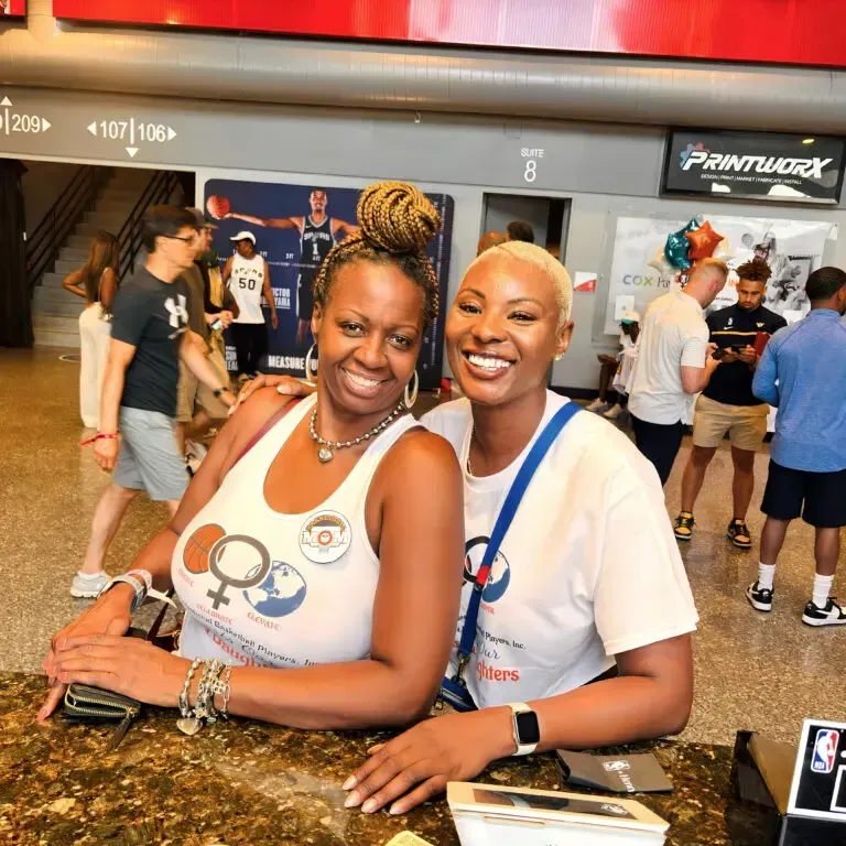 Two smiling women pose together indoors, wearing white shirts with logo.