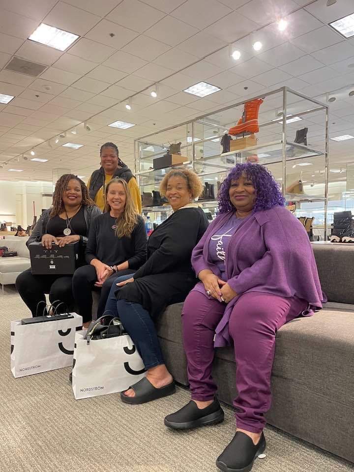 Six women pose in a shoe store. They are sitting on a bench, some holding shopping bags.