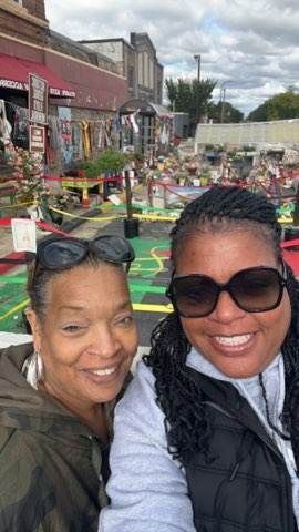 Two smiling women pose outdoors with a memorial in the background.