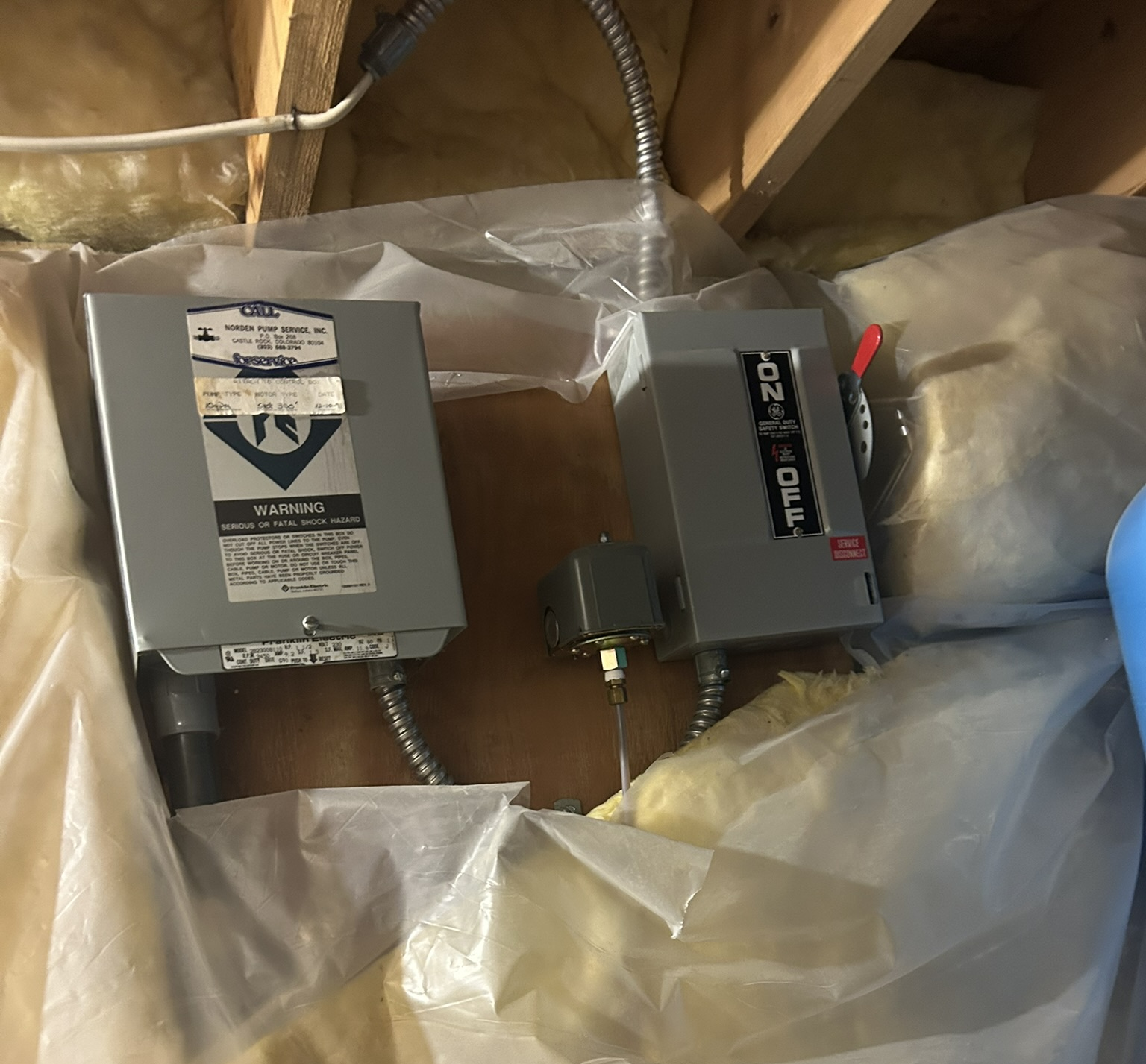 Two gray electrical boxes with wires mounted among exposed wooden attic rafters and insulation plastic.