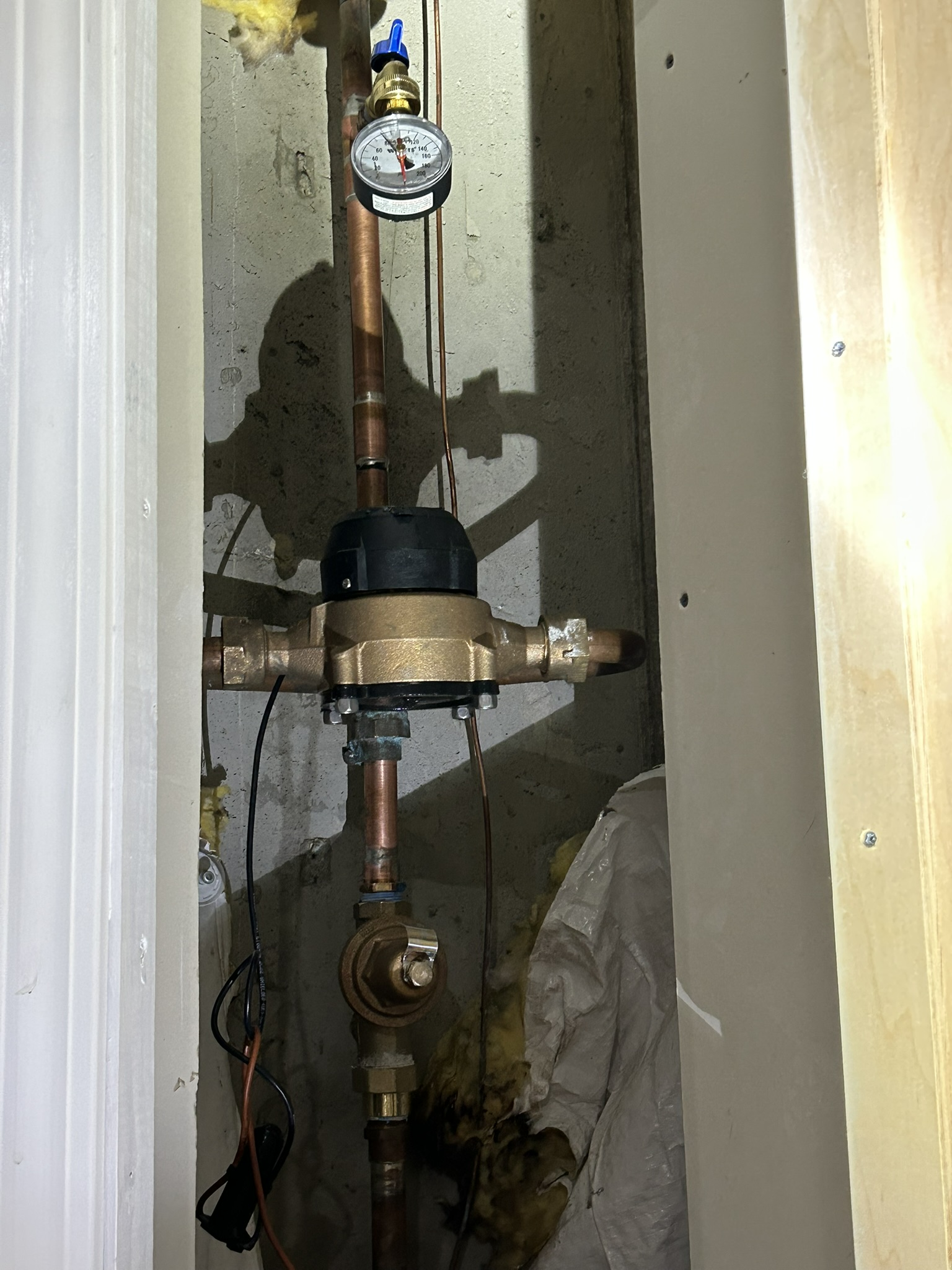 Vertical plumbing pipes with a pressure gauge and valve in a narrow utility alcove.