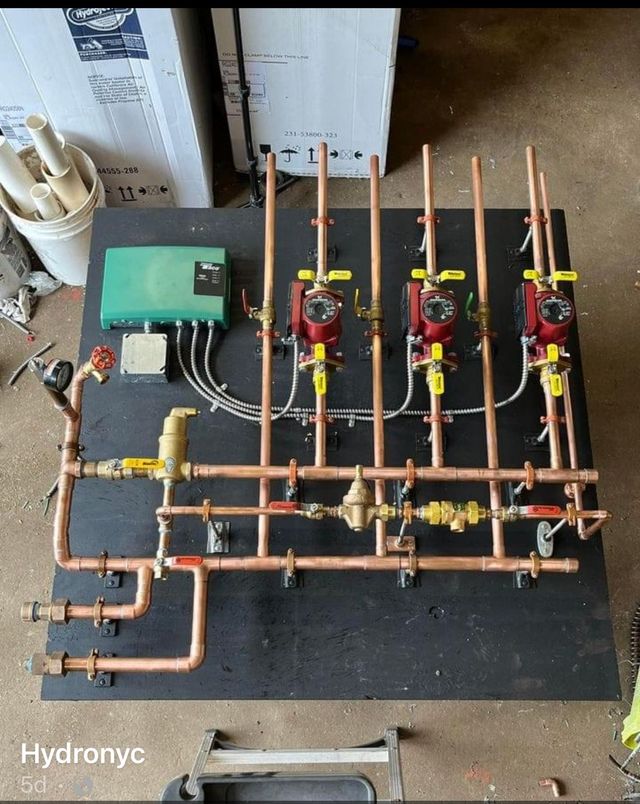 Copper hydronic plumbing manifold with three red pumps and green control box on a black board
