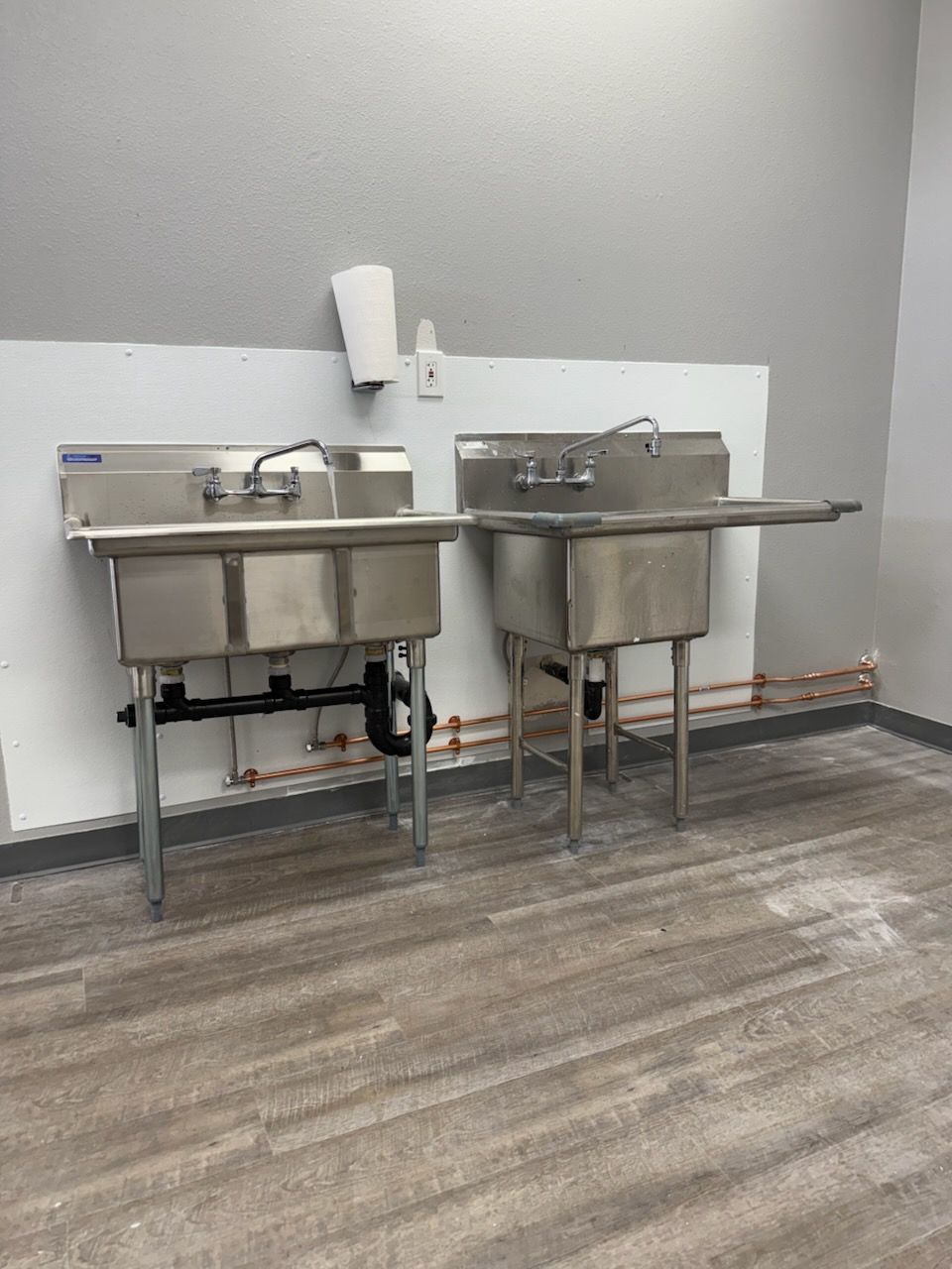 Two stainless steel sinks with faucets against a white wall in a room with wood-look flooring