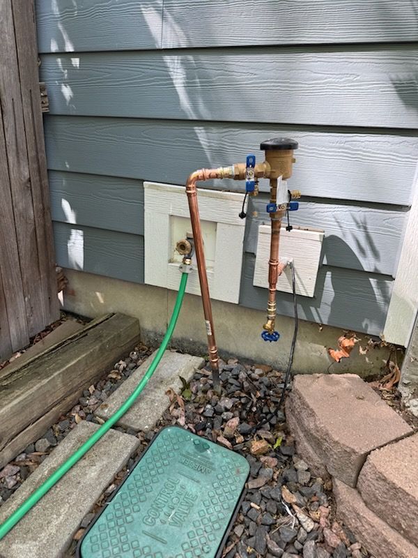 Outdoor water meter and shutoff valve beside a house, with green hose and gravel bed.