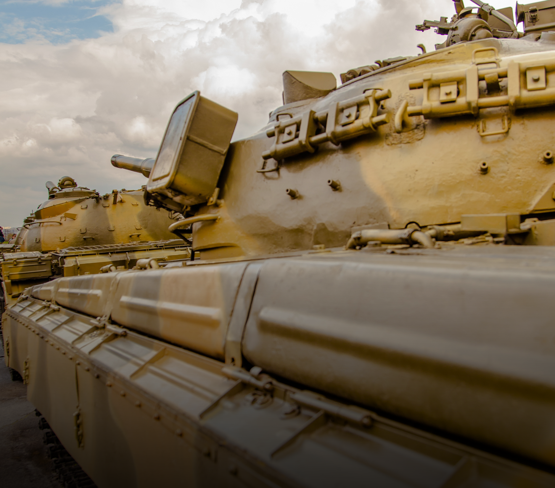 Tanks in desert camouflage parked outdoors under a cloudy sky.