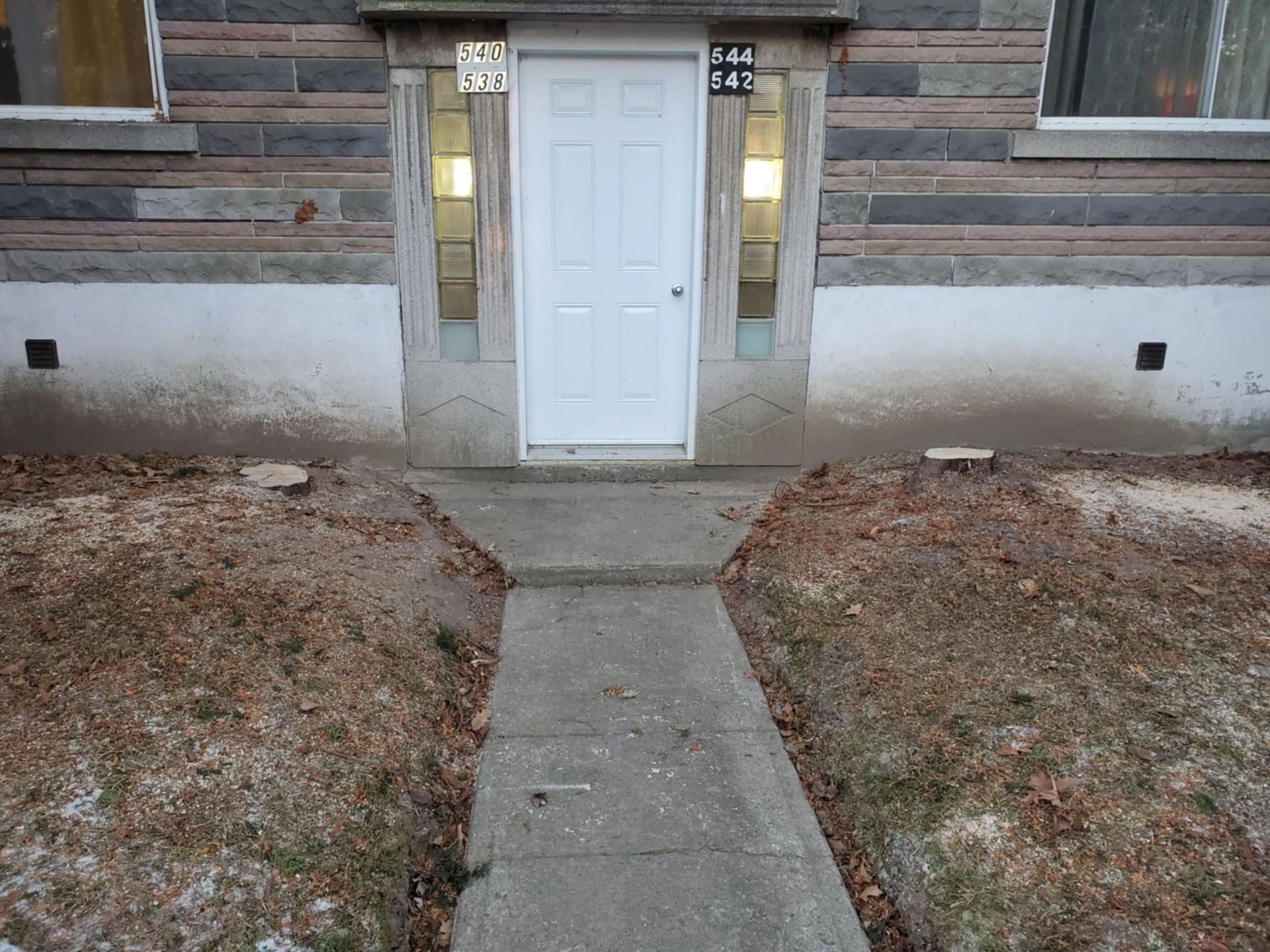 Allée en béton menant à une porte blanche dans un bâtiment à l'extérieur en pierre et en stuc peint.