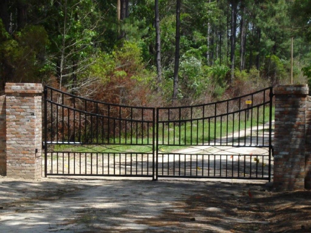 A wrought iron gate is open to a dirt road surrounded by trees.