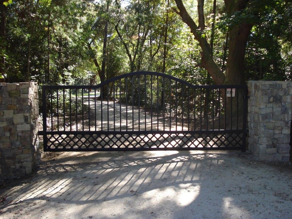 A black gate is open to a driveway surrounded by trees