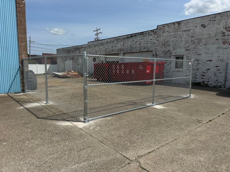 Chain-link fenced enclosure beside a red dumpster against a brick building on a concrete lot
