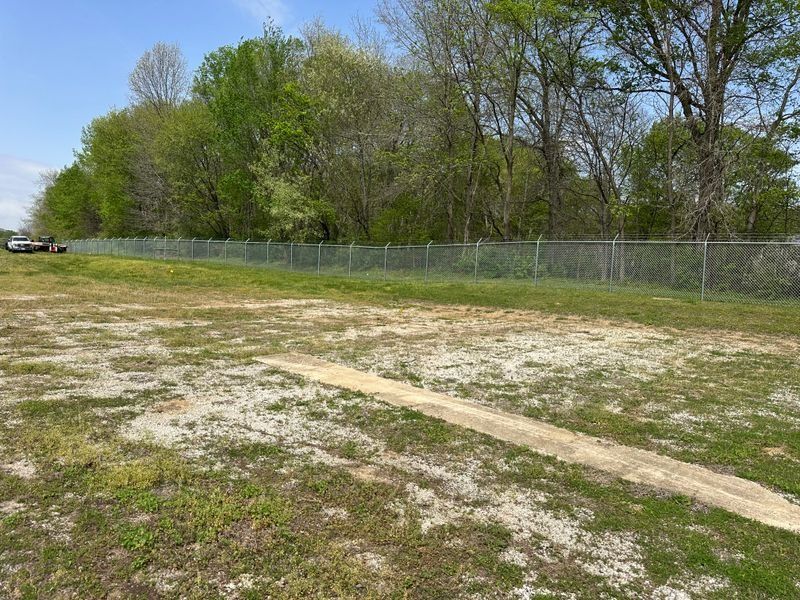 Grassy field with a dirt path beside a chain-link fence and trees under a clear sky