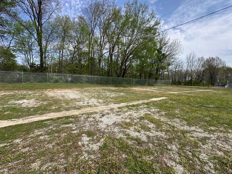 Open grassy field with patchy dirt, chain-link fence, and trees under a partly cloudy sky