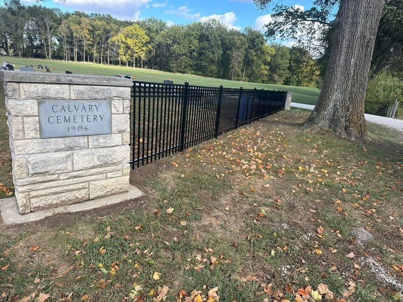 Calvary Cemetery entrance sign beside a black fence, grass, and a large tree in a sunny park setting