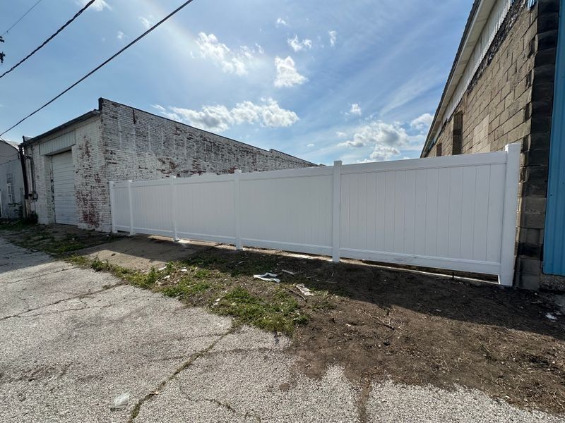 White vinyl fence beside a weathered concrete building on a cracked alley under a blue sky