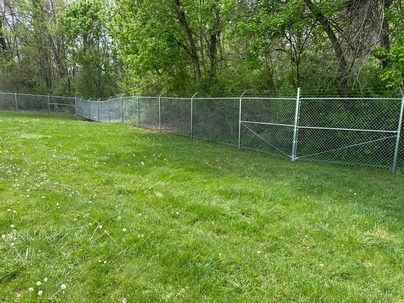 Chain-link fence along a grassy field beside leafy trees and a wooded area.