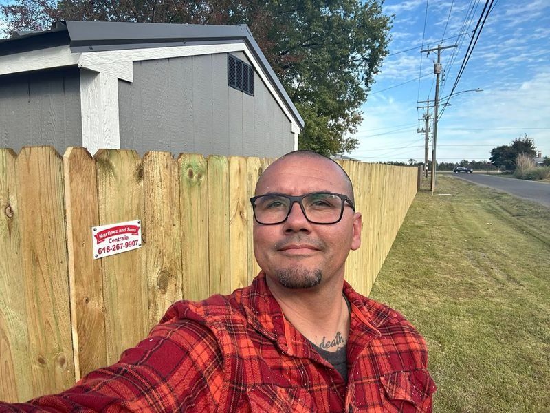 Man taking a selfie beside a wooden fence on a sunny roadside, wearing a red plaid shirt and glasses.